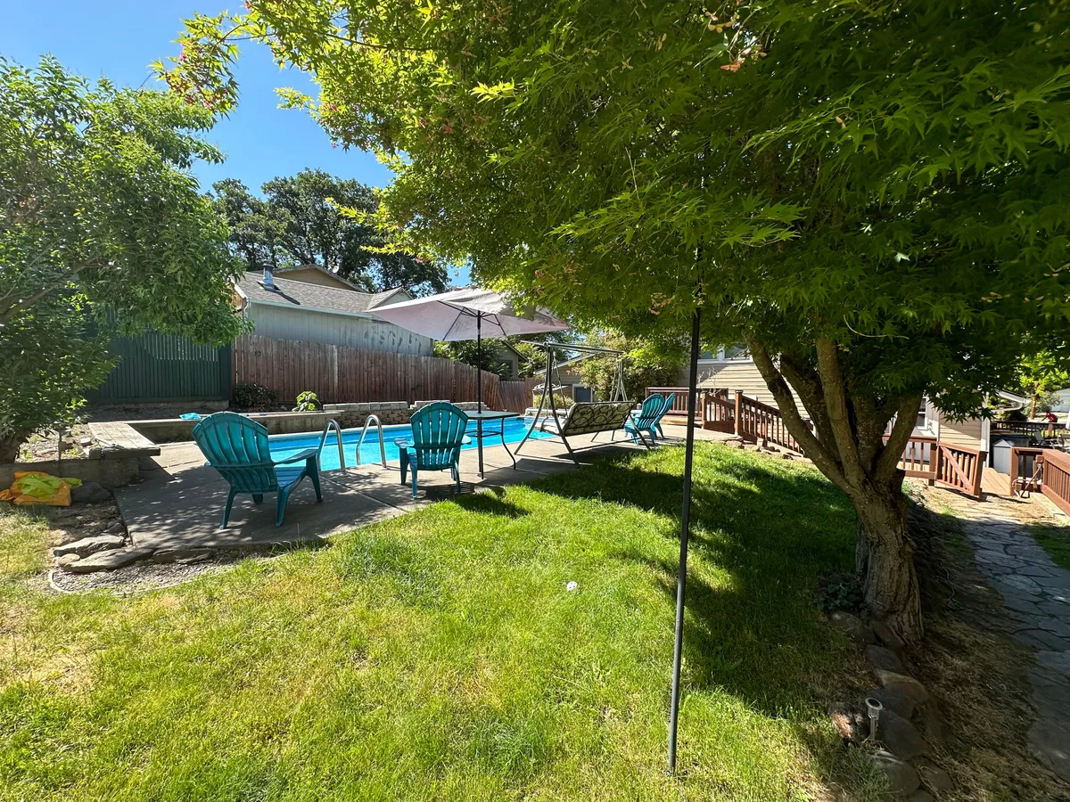 An exterior scene featuring a sparkling swimming pool, lounge chairs, a patio umbrella, green grass, and trees. The setting feels serene and welcoming, perfect for unwinding or socializing after a day of exploration.