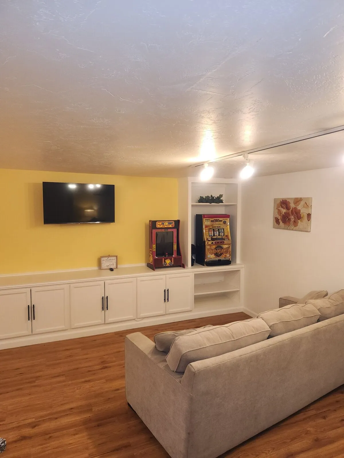 A stylish living area featuring a comfortable sofa facing a wall-mounted TV, with decorative artwork and playful arcade machines nearby. The space balances relaxation and entertainment, with cozy seating inviting you to rest and modern décor providing a soothing backdrop.