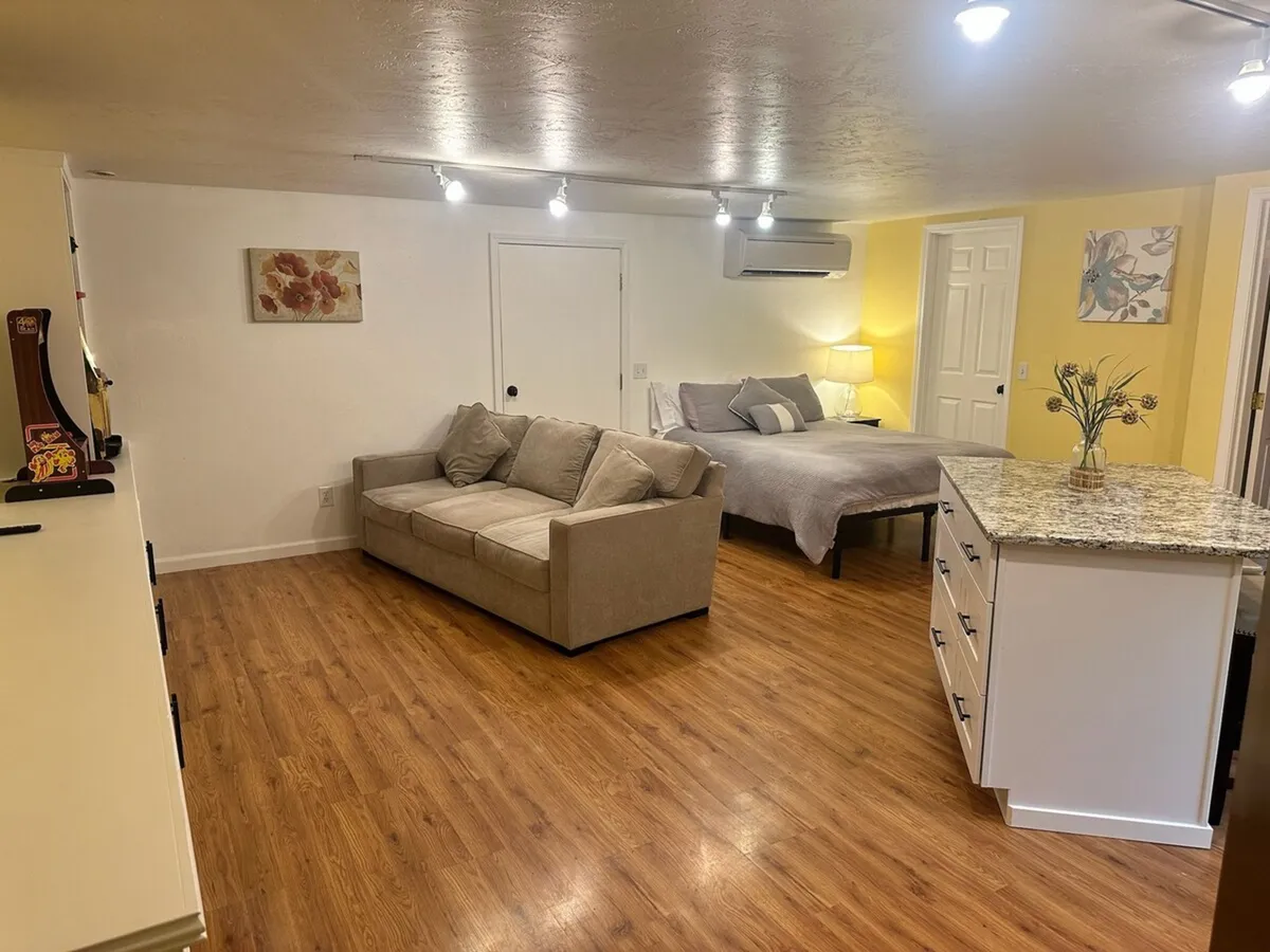 This image showcases a cozy, inviting interior with a sofa and bed in view, complemented by a kitchen island in the background. The main area is bathed in soft lighting, emphasizing plush seating and a seamless, open layout. The scene evokes relaxation, with hints of someone reading or unwinding, suggesting a tranquil space ideal for downtime.
