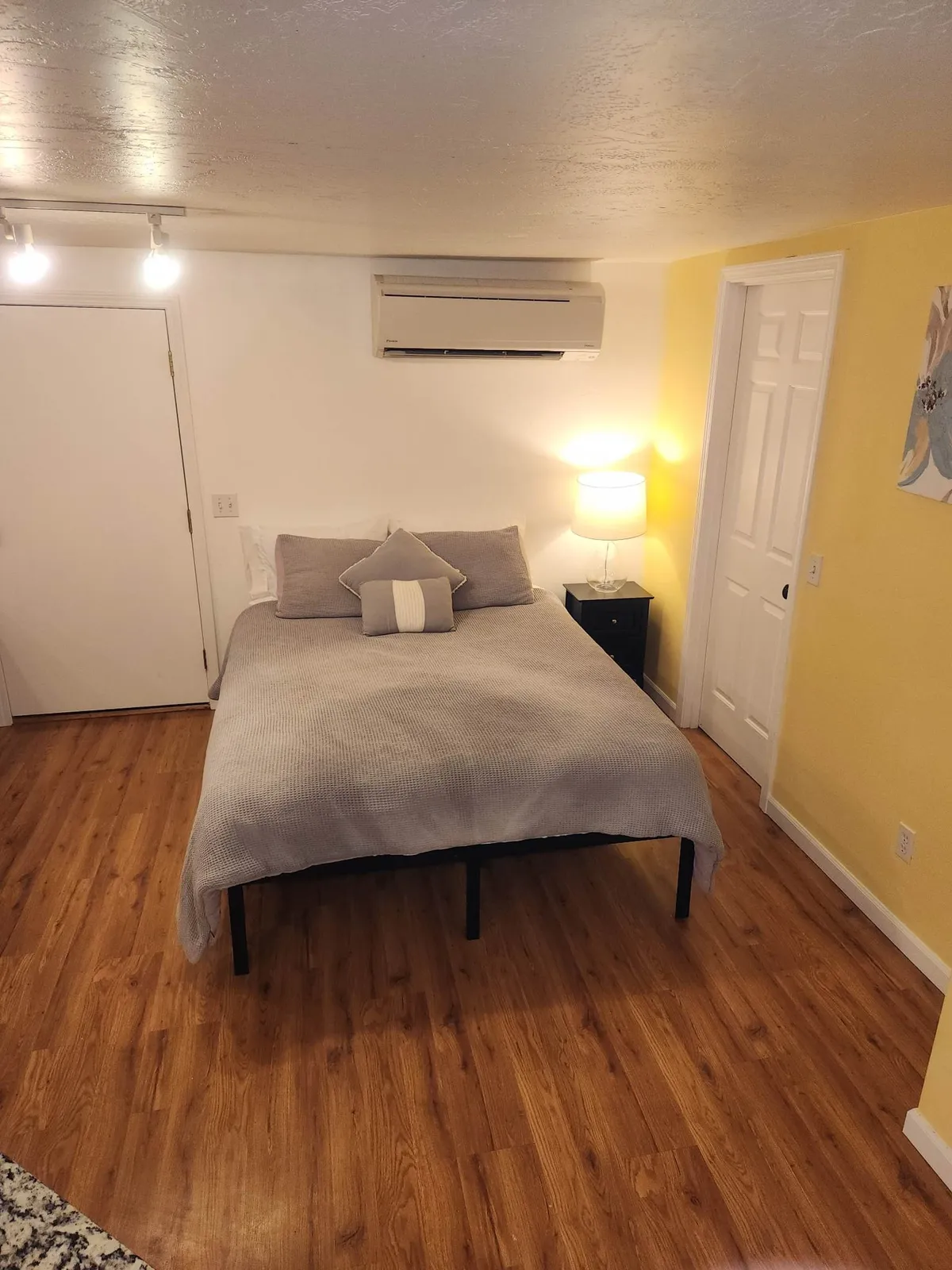 This image features a cozy bedroom with a plush bed as the central focus. Soft linens, inviting pillows, a bedside lamp, and calming artwork on the wall all contribute to a serene and restful atmosphere. Morning light filters gently through the window, casting a warm glow across the room.