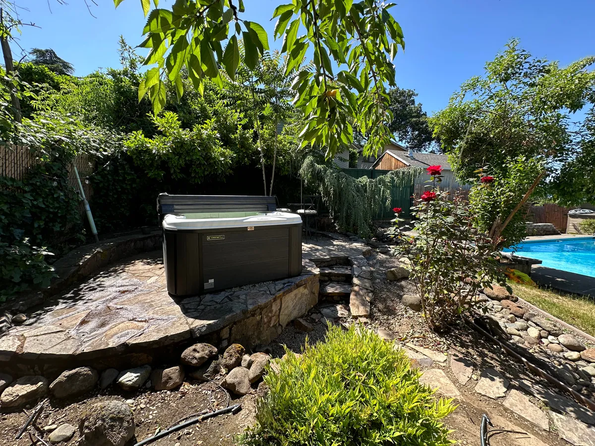 A peaceful exterior image with a hot tub nestled among flowering plants, a stone pathway, and a view of the swimming pool. The scene exudes tranquility and relaxation, with touches of nature enhancing the outdoor experience.
