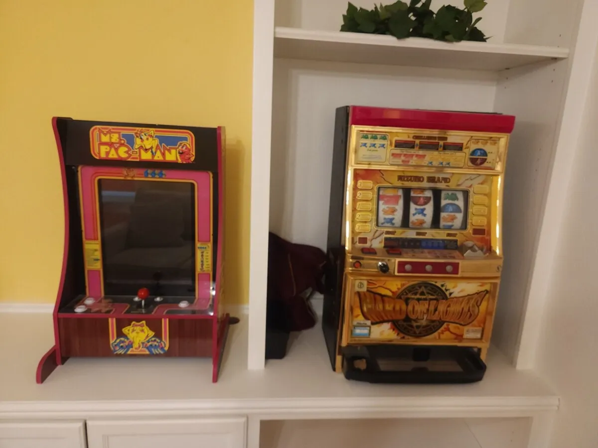This image captures a vibrant, retro-themed interior space featuring a Ms. Pac-Man arcade machine and a slot machine. The atmosphere is lighthearted, with people enjoying classic games and socializing, evoking a sense of fun and communal energy.