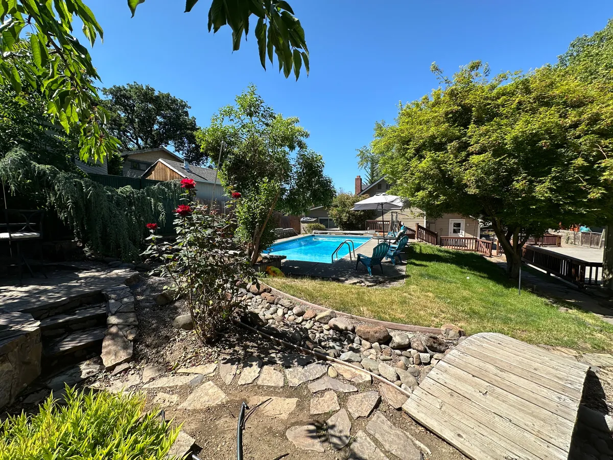 This exterior image showcases a swimming pool surrounded by lush greenery, a stone pathway, colorful flowers, and a deck with chairs, all set under a relaxing sky. The inviting scene suggests a seamless connection to nature, with spaces for swimming, sunbathing, and gathering outdoors.