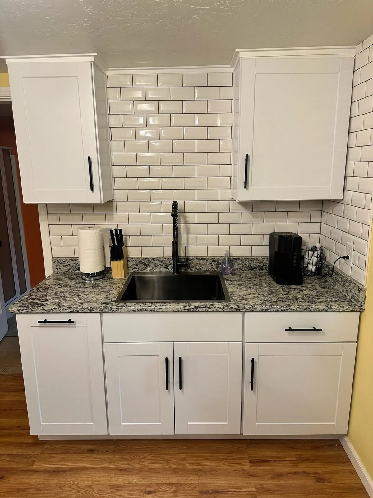 The image showcases a modern, inviting kitchen area featuring gleaming granite countertops, a sleek black faucet, crisp white cabinetry, and a classic subway tile backsplash. The setup feels open and functional, perfect for preparing meals or gathering with loved ones.