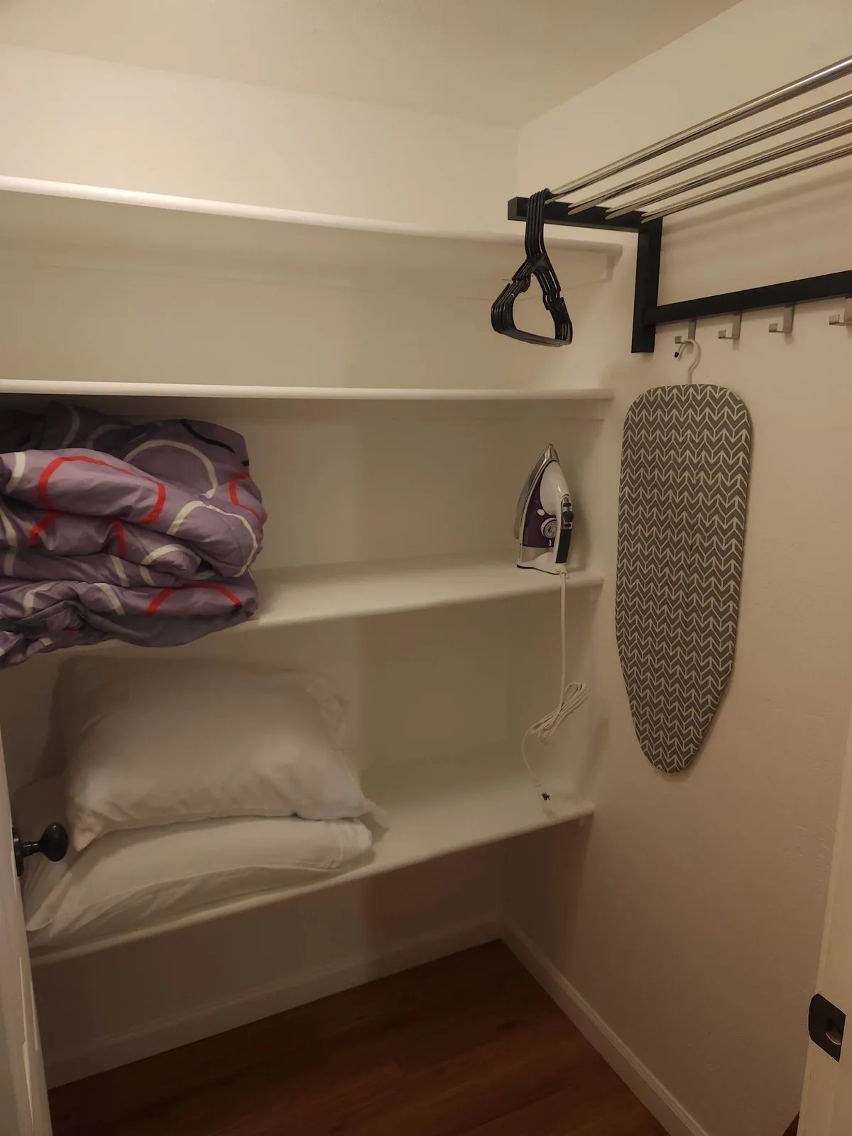 This image features neatly arranged shelves stocked with household essentials like an iron, ironing board, folded blankets, and pillows. The space feels organized and practical, suggesting readiness for guests to settle in and feel at home.