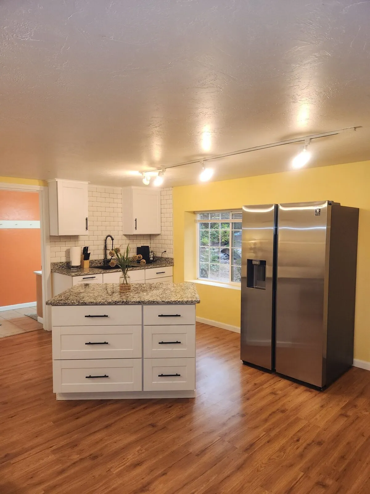 A bright, cheerful kitchenette with a granite countertop island, stainless steel refrigerator, a vibrant yellow wall, and ample natural light streaming in from the window. The setting is welcoming and encourages both cooking and social interaction.