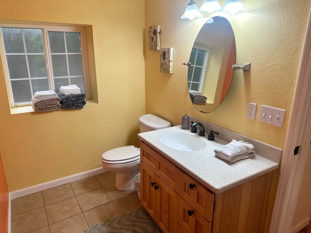 A modern bathroom with a wooden vanity, a round mirror, neatly stacked towels, and a subtle pop of color from a yellow wall. A window brings in natural light, enhancing the tranquil, refreshed feeling of the space.